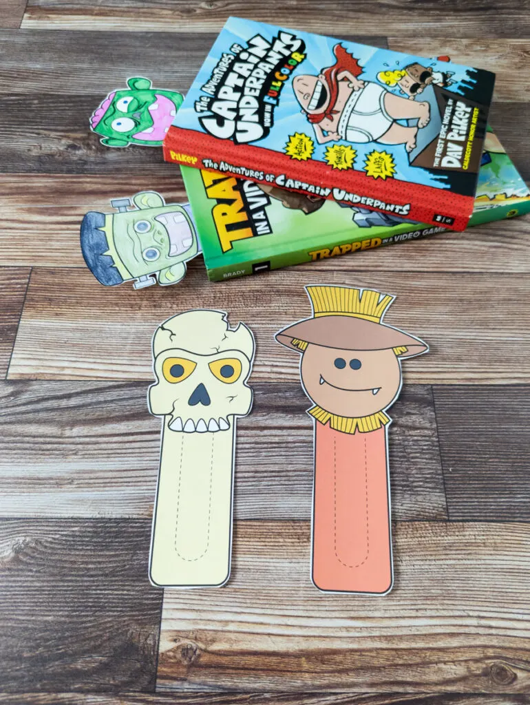 Full color skeleton and scarecrow bookmarks cut out and laying side by side. Zombie and Frankenstein's monster bookmarks colored in and sticking out of two hard cover graphic novels for kids.