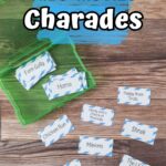 Overhead view of assorted charades cards with kid movie titles cut out and laying on table. Some cards are in an open green task box. Top of the photo has white text on a blue splash of color that says Kids' Movies Charades.