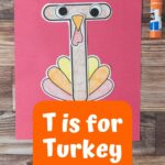 Paper pieces assembled to create a turkey and using a T shape as the body on a piece of red construction paper. White text on orange near bottom says T is For Turkey Craft.
