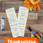 Two strips of printed out word cards for Thanksgiving Charades next to a blue sand timer and a pair of scissors. A small pumpkin and fall leaves decorate the top corner.