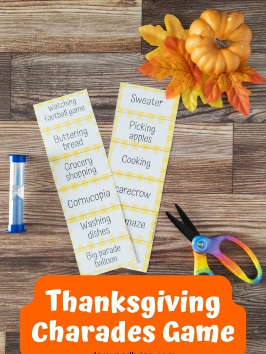 Two strips of printed out word cards for Thanksgiving Charades next to a blue sand timer and a pair of scissors. A small pumpkin and fall leaves decorate the top corner.