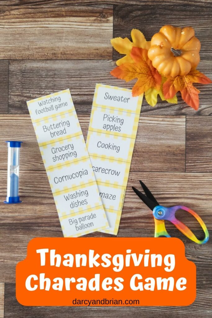 Two strips of printed out word cards for Thanksgiving Charades next to a blue sand timer and a pair of scissors. A small pumpkin and fall leaves decorate the top corner.
