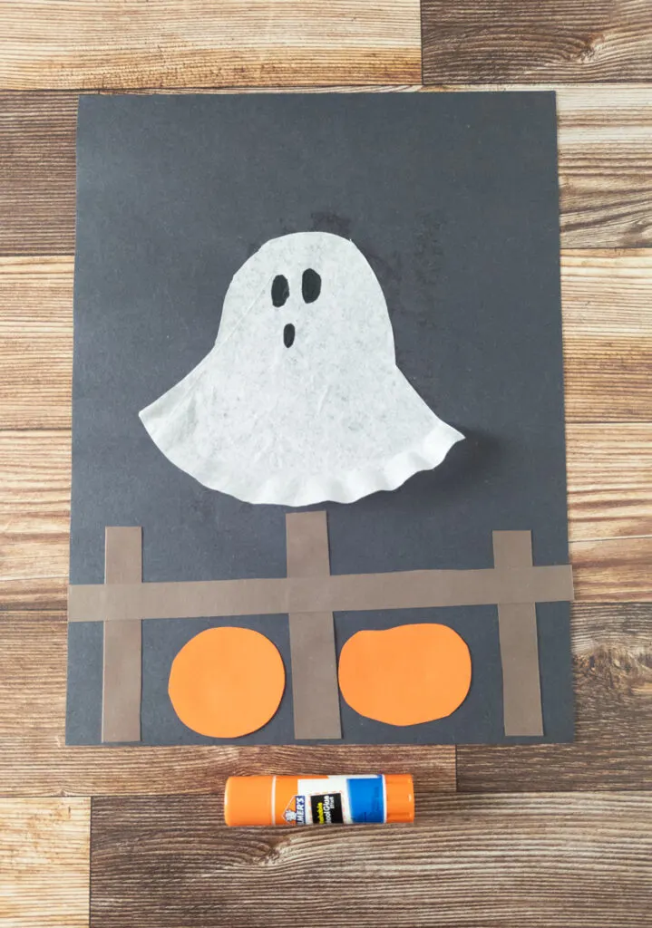 Coffee filter ghost glued to black paper and floating above a fence and pumpkins made out of construction paper.
