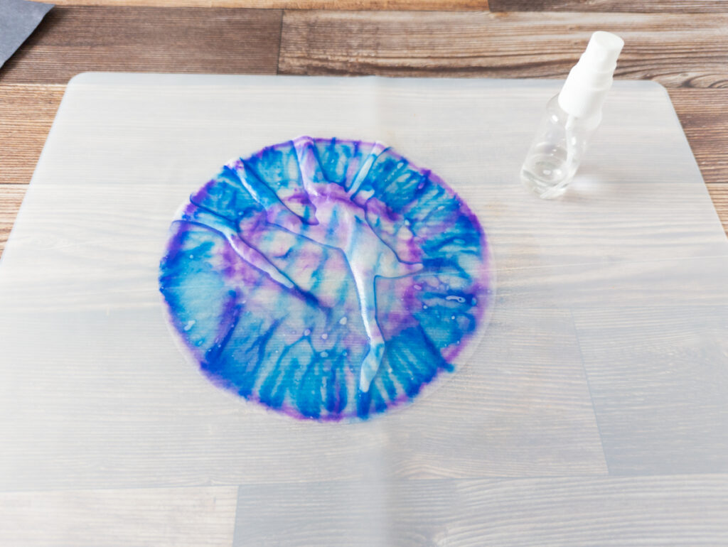 Blue and purple colored coffee filter sprayed with water laying on a translucent craft mat. A small fingertip spray bottle stands next to it.