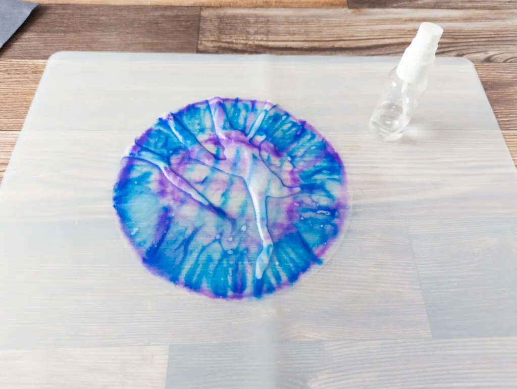 Blue and purple colored coffee filter sprayed with water laying on a translucent craft mat. A small fingertip spray bottle stands next to it.