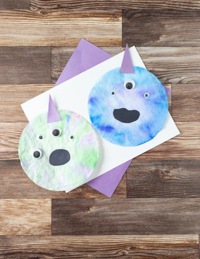 Two finished coffee filter monster kid crafts laying on top of white and purple paper. One monster is greenish and the other is bluish. They have multiple googly eyes.