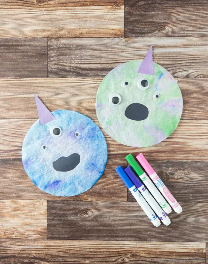 Blue colored coffee filter monster and green tie dyed coffee filter laying next to each other with purple paper horns and multiple googly eyes. Several markers lay next to them.