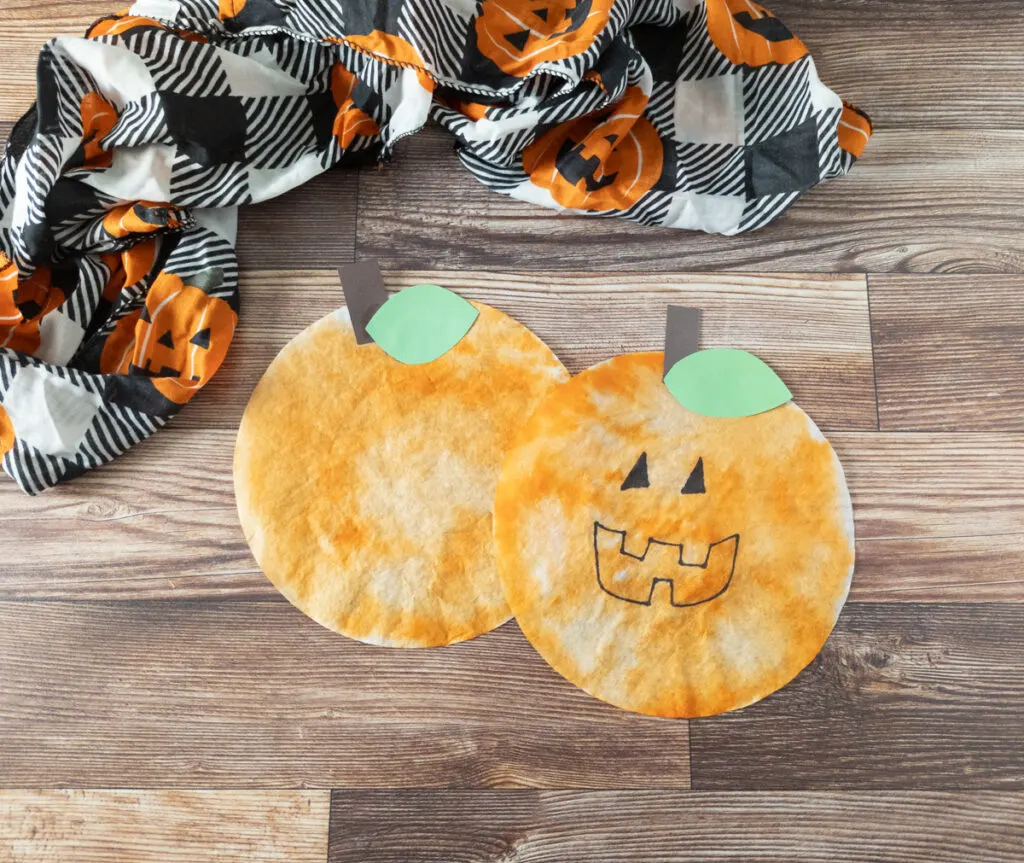Two pumpkins made using coffee filters laying next to each other. A decorative piece of fabric with jack-o-lanterns on it is along the top part of the photo.