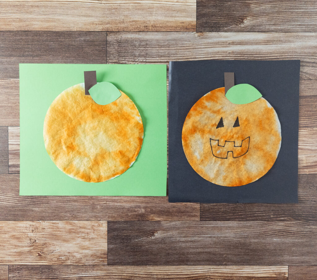 Two finished coffee filter pumpkin crafts. One without a face is on light green paper. One with a face drawn on is on black paper.