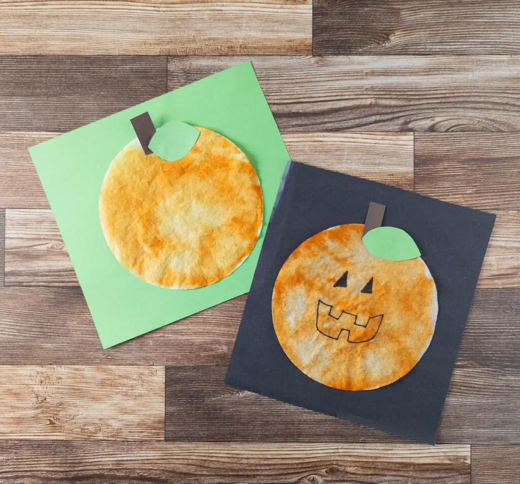 Two different coffee filter pumpkins. One with a face on black paper. One plain pumpkin on green paper. Both are laying tilted near each other.