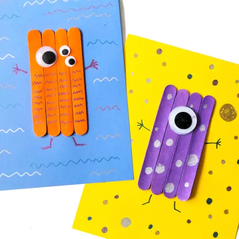 Orange craft stick monster with multiple eyes on blue paper overlapping yellow paper with purple one eyed monster on it.
