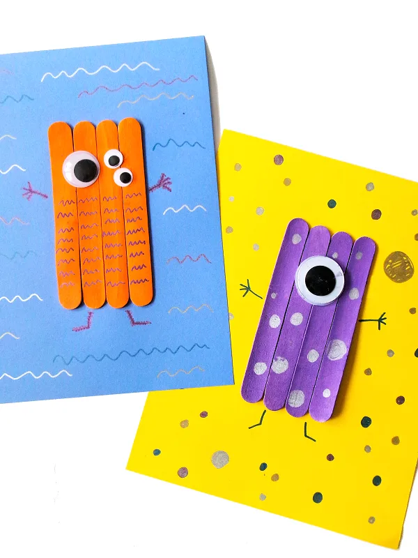 Orange craft stick monster with multiple eyes on blue paper overlapping yellow paper with purple one eyed monster on it.