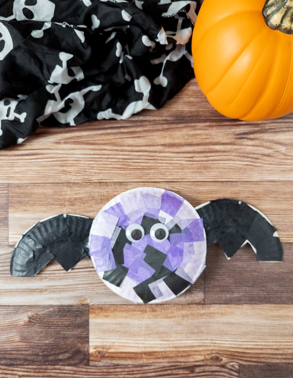 Top down view of finished bat craft made with tissue paper and paper plates. The craft is laying near an orange pumpkin decoration and a black cloth with white skulls on it.
