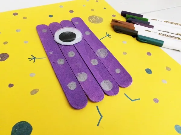 Purple popsicle stick monster with one large googly eye glued on yellow paper. Metallic paint markers used to draw stick arms and legs and decorate the monster's body with dots.