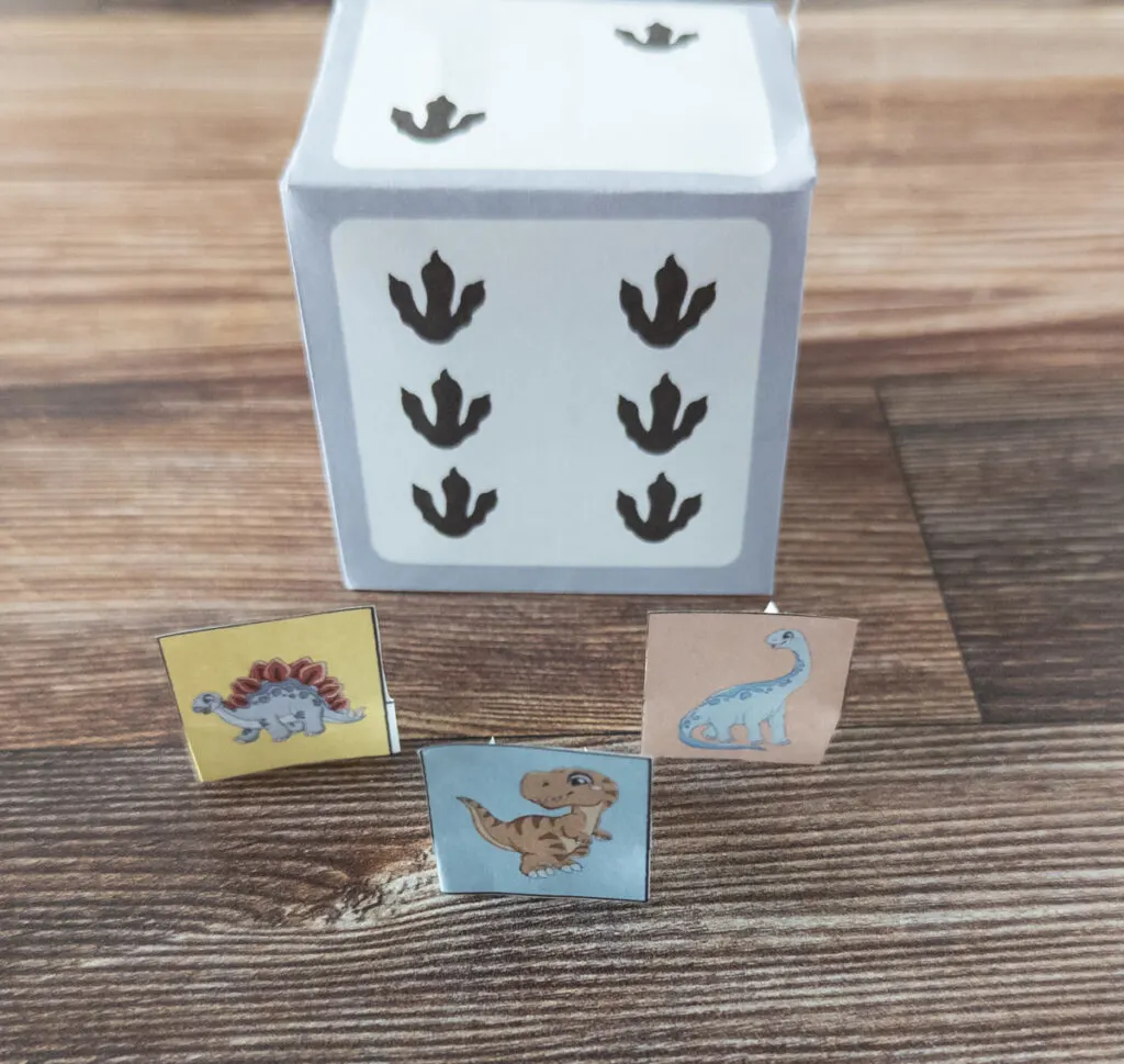 Close up of three different dinosaur player tokens cut out and standing up in front of custom paper dice that uses dino tracks as pips.