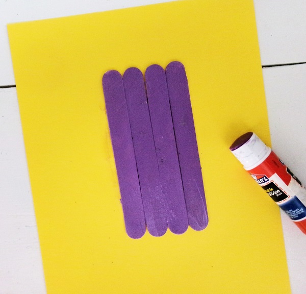 Four craft sticks painted purple and glued to yellow cardstock. Open glue stick lays next to it.