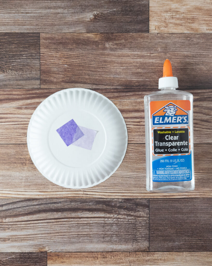 A couple squares of purple tissue paper glued to the middle of a small paper plate. Next to the plate is a bottle of clear glue.