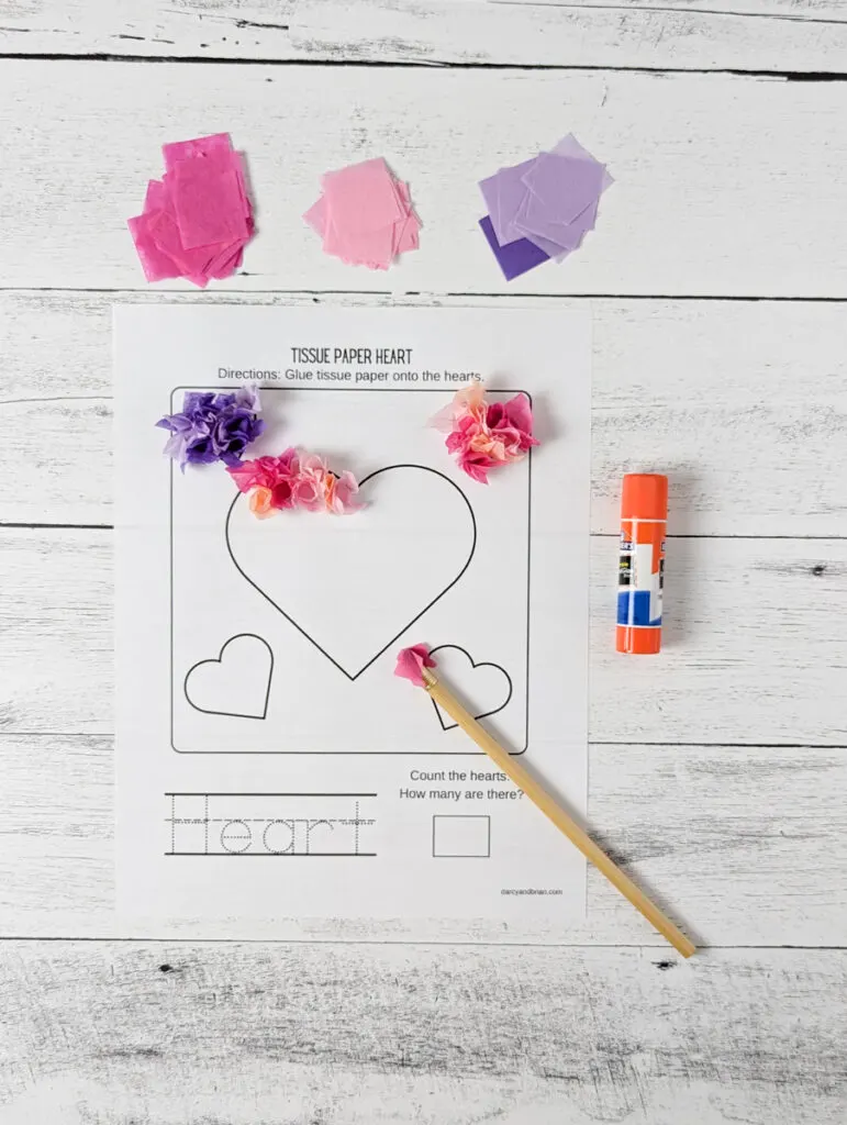 Piles of different colored tissue paper and a glue stick lay around heart craftivity worksheet. Some pieces of tissue have been glued down. A pencil with paper wrapped around one end lays diagonally across the template.