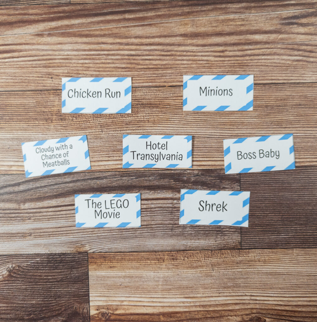 Seven movie charades for kids prompt cards cut out and arranged on the table. The include movie titles Chicken Run, Minions, Cloudy With a Chance of Meatballs, Hotel Transylvania, Boss Baby, The LEGO Movie, and Shrek.
