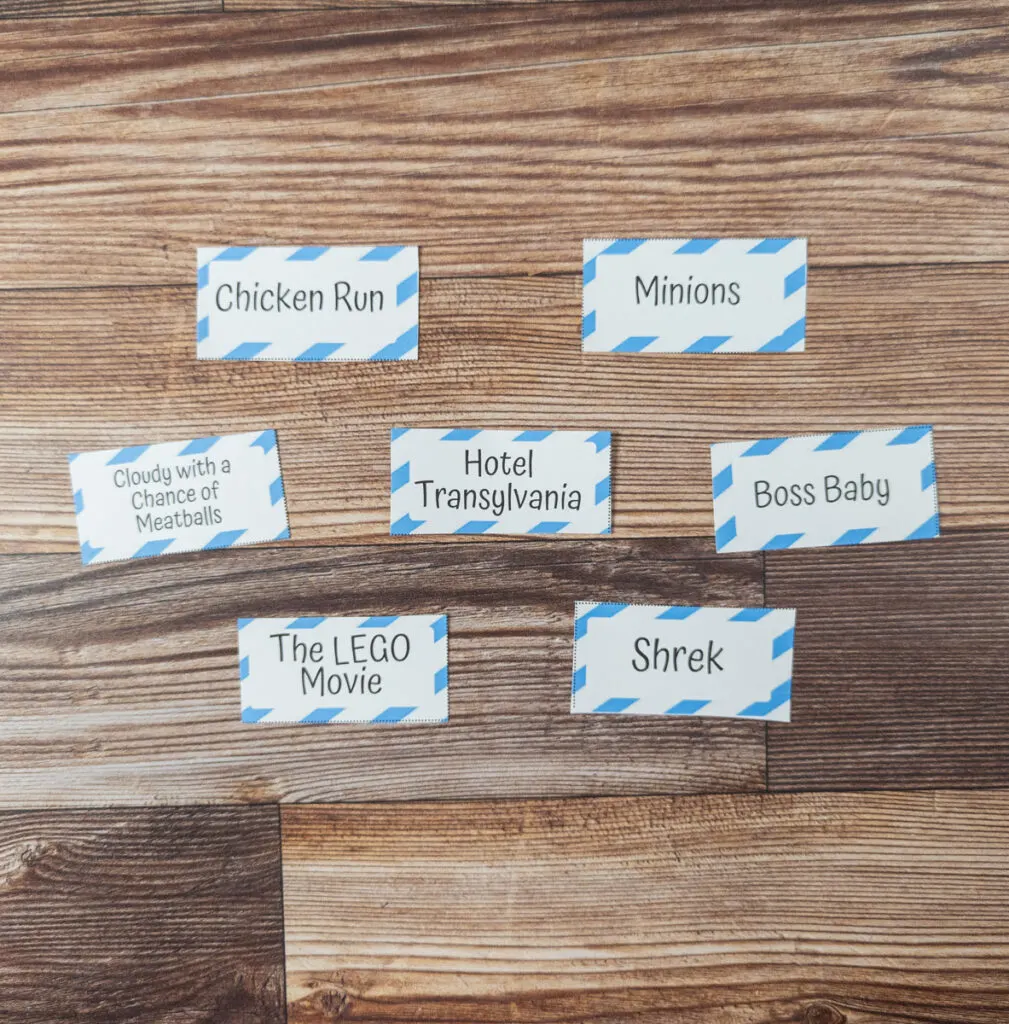 Seven movie charades for kids prompt cards cut out and arranged on the table. The include movie titles Chicken Run, Minions, Cloudy With a Chance of Meatballs, Hotel Transylvania, Boss Baby, The LEGO Movie, and Shrek.