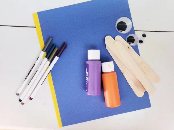 Yellow and blue cardstock, purple and orange paint, popsicle sticks, googly eyes, and metallic paint markers lay on table.