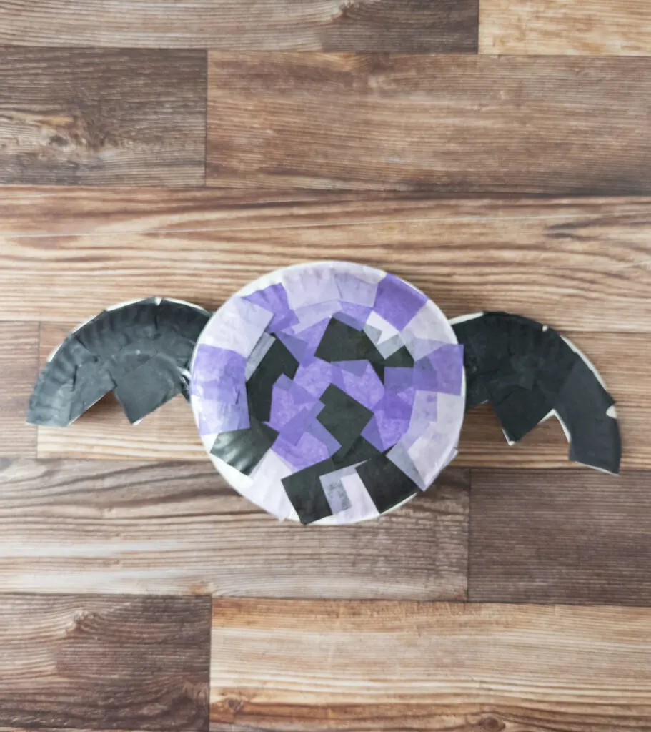 Top down view of round paper plate covered in purple and black tissue paper squares and paper plate wings covered in black paper.