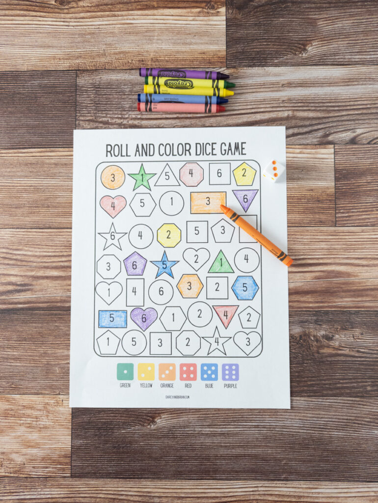 Roll and color dice game with shapes to color in. Crayons along the top and an orange crayon laying on the paper next to a die with the number three rolled. 