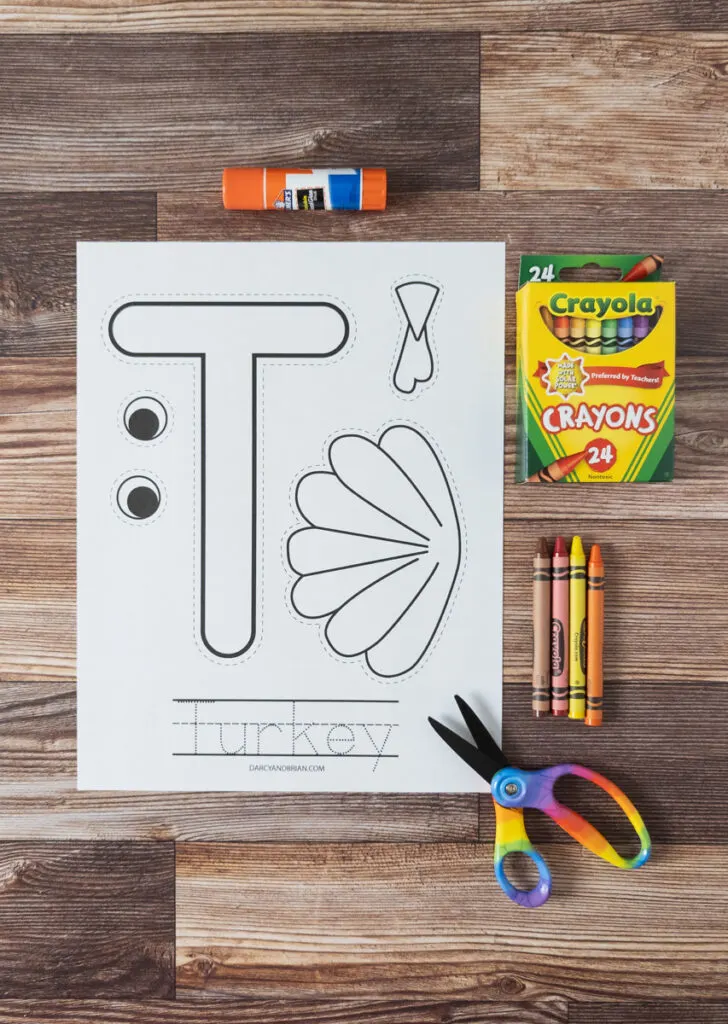 Top down view of printed out t is for turkey craft template, glue stick, crayons, and scissors on table.
