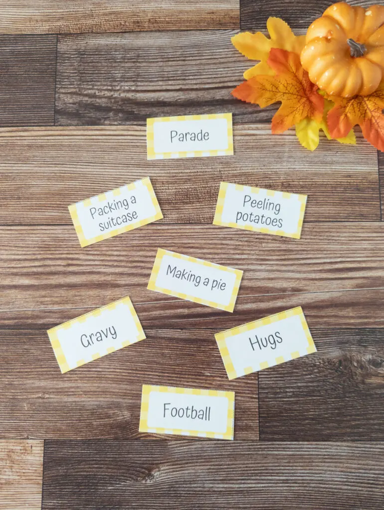 Seven charades cards with a yellow gingham border arranged on table near pumpkin and leaf props. Cards say: parade, packing a suitcase, peeling potatoes, making a pie, gravy, hugs, and football.