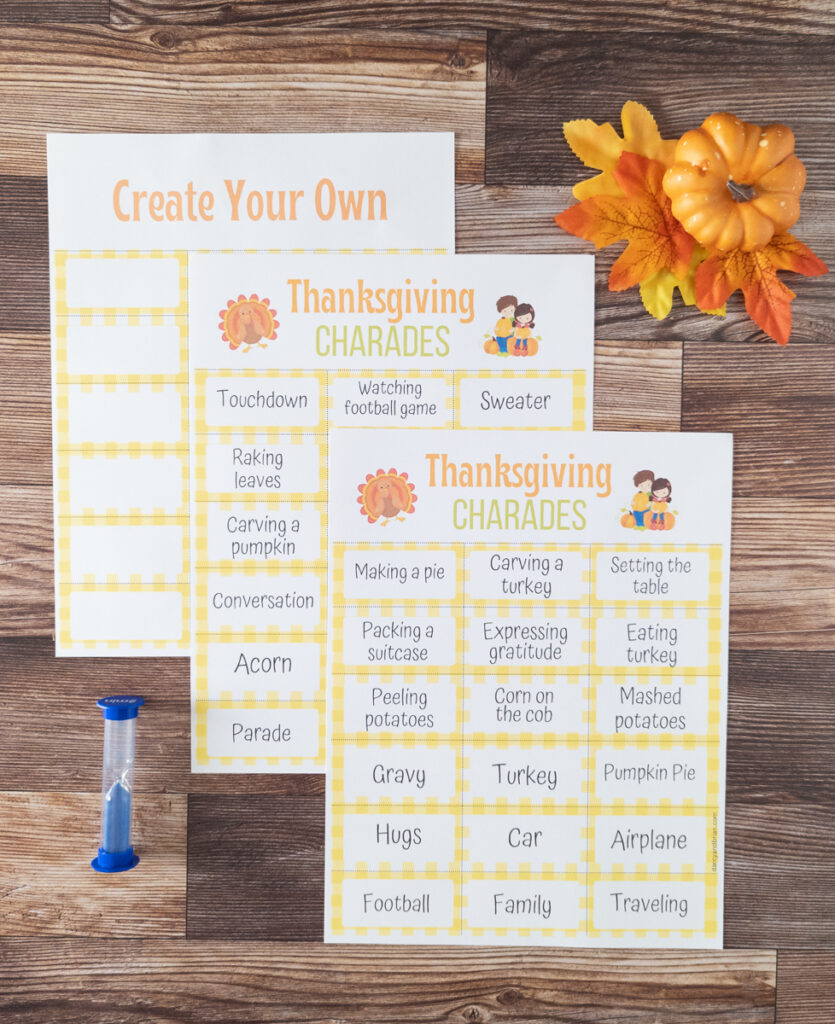 Three pages of Thanksgiving charades game printed out and overlapping diagonally on table. Blue sand timer in lower left corner and pumpkin and leaf props in upper right corner.