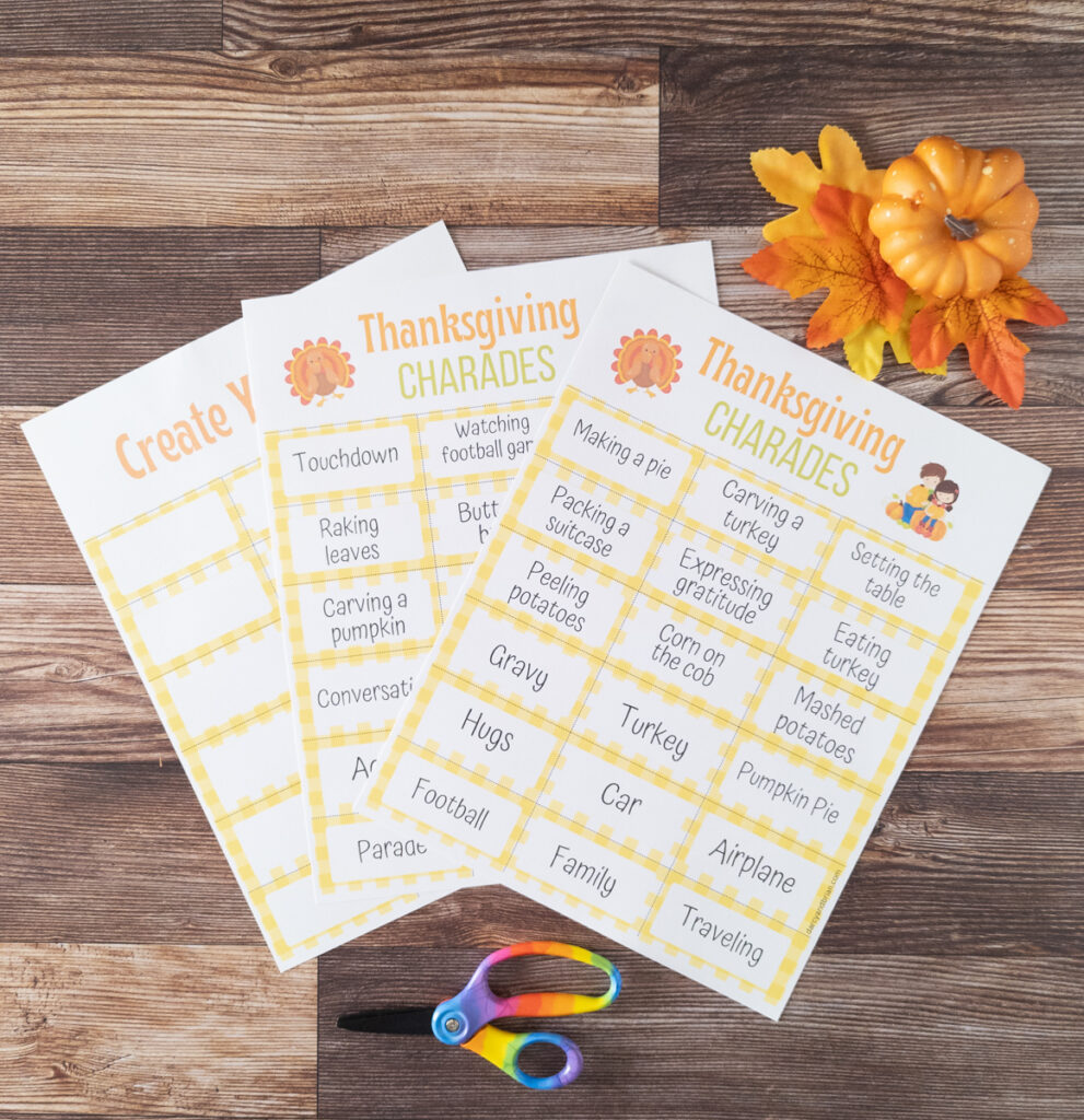 Two pages of Thanksgiving themed charades cards and one page of blank cards fanned out on table next to scissors and a small pumpkin and leaves.