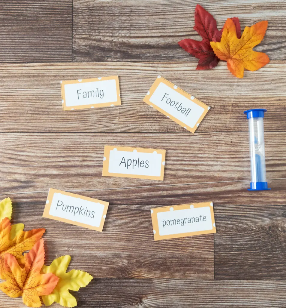 Five Pictionary word cards cut out and laying on table next to blue sand timer. Faux fall leaves decorate the corners of the photo. The word cards say: family, football, apples, pumpkins, and pomegranate.