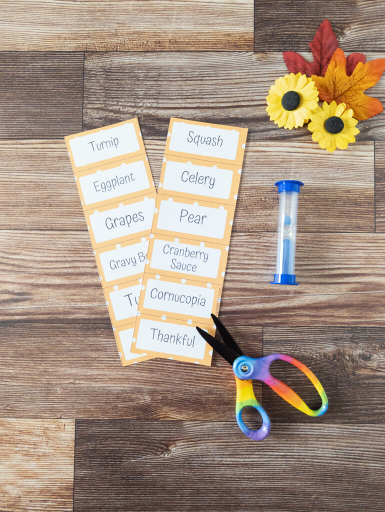 Two columns of Pictionary word cards overlapping each other on table next to scissors and blue sand timer.