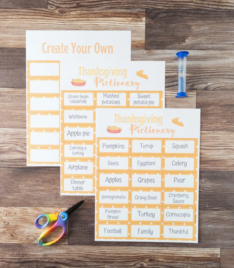 Three pages of Thanksgiving Pictionary printed out and overlapping each other on table. Two pages have word card prompts and one back is blank cards for creating your own. Blue sand timer and scissors lay next to pages.