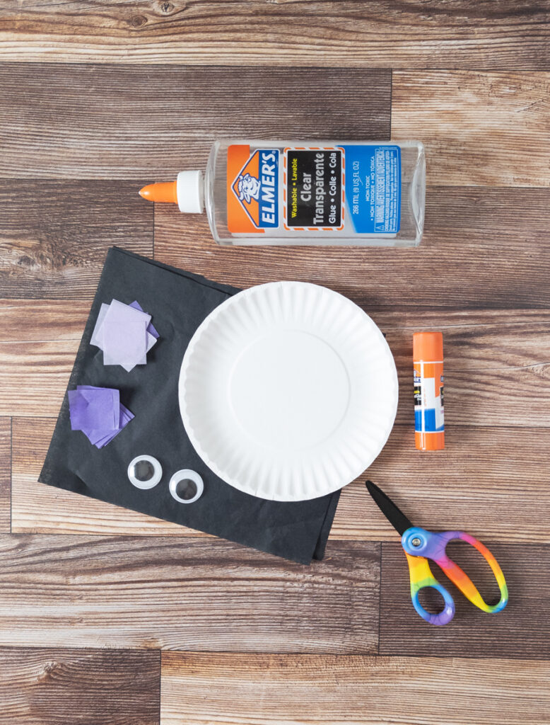 Overhead view of sheets of black tissue paper, squares of purple tissue paper, googly eyes, small paper plates, scissors, glue stick, and a bottle of clear glue.