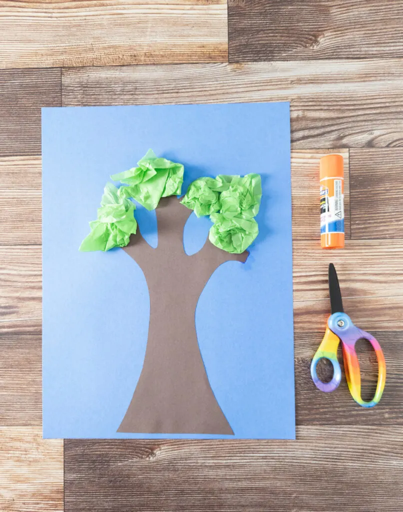 Tissue paper tree glued on blue paper. Glue stick and scissors lay next to the project.