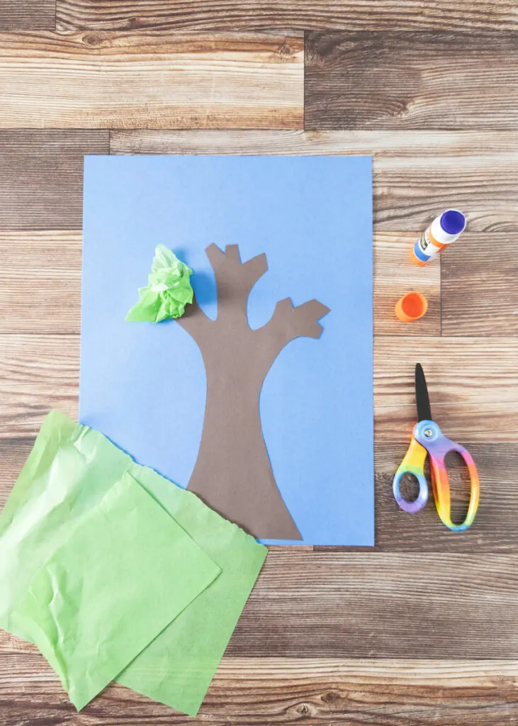 Gluing pieces of green tissue paper around brown paper tree branch.