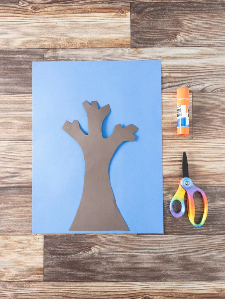 Tree trunk and branches cut out of brown paper and placed on blue paper. Scissors and glue stick lay on the right.