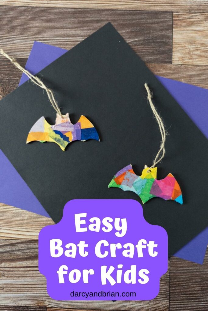 Two wooden bat shapes covered in colorful tissue paper with twine to hang them. They are laying on top of black and purple construction paper.