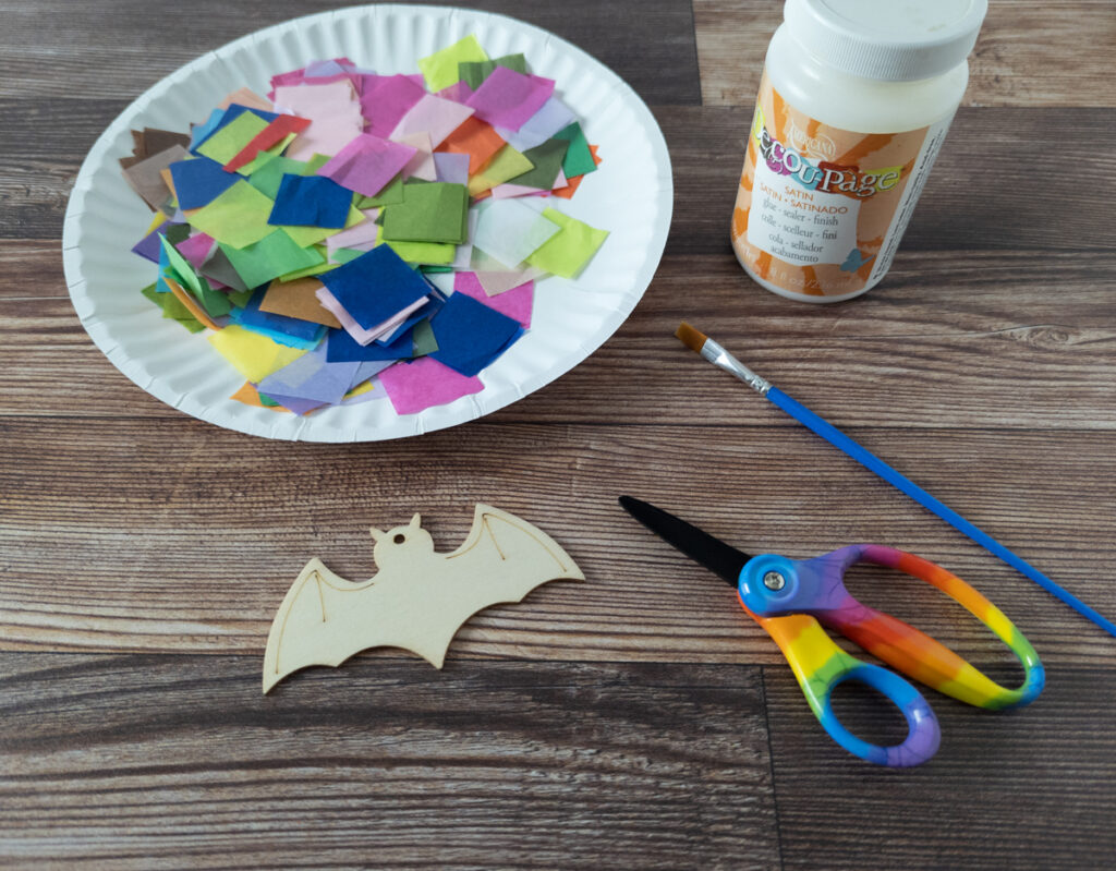 Assorted colors of square tissue paper on a paper plate, a flat wooden bat, scissors, decoupage glue, and small paintbrush.