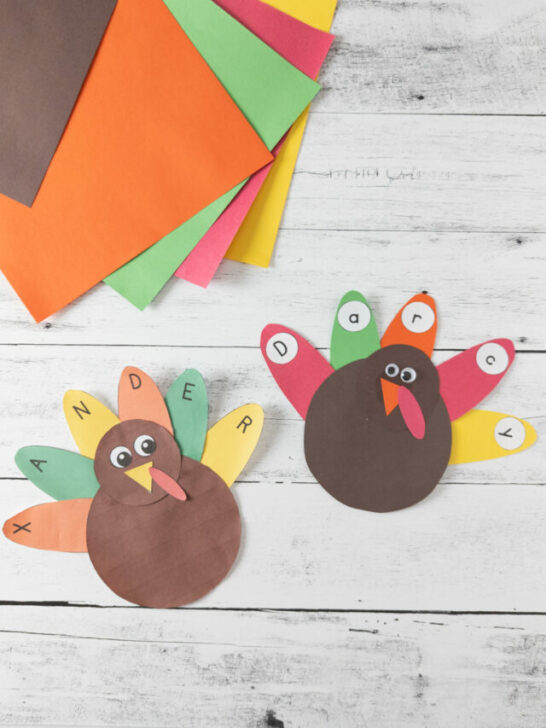 Two finished turkey crafts with the feathers spelling a name. Assorted construction paper is fanned out in upper left corner.