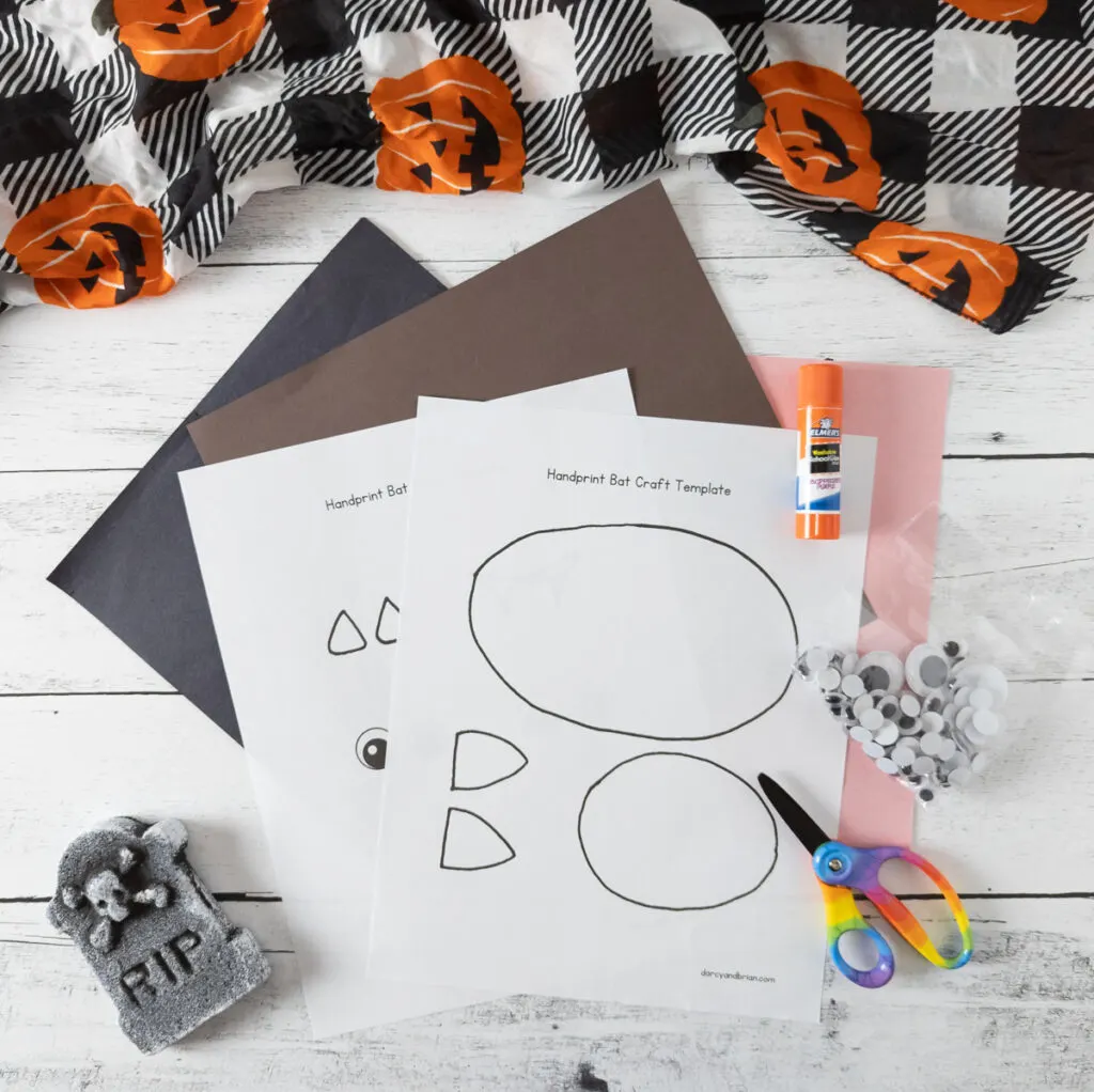 Bat craft template, construction paper in black, brown, and pink, glue stick, scissors, and googly eyes laying out. Also fabric with black and white plaid and jack-o-lanterns along the top. A small prop headstone in the lower left corner.