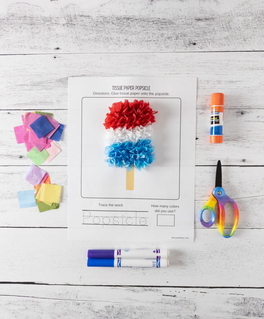 Overhead view of completed red, white, and blue tissue paper popsicle. Paper is sticking up creating a shaggy textured look. Assorted colors of paper, markers, scissors, and glue stick surround the project.