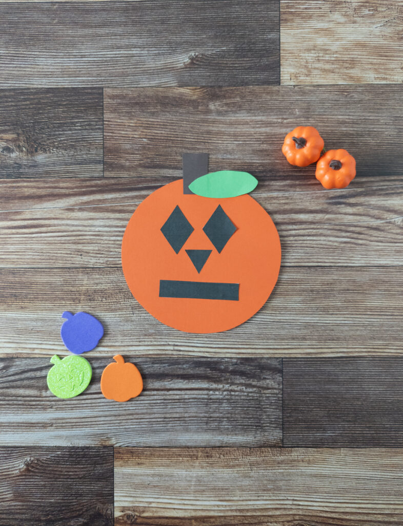 Completed shape pumpkin craft made using orange, brown, and green construction paper. Black shapes used to make the face were printed out on paper.