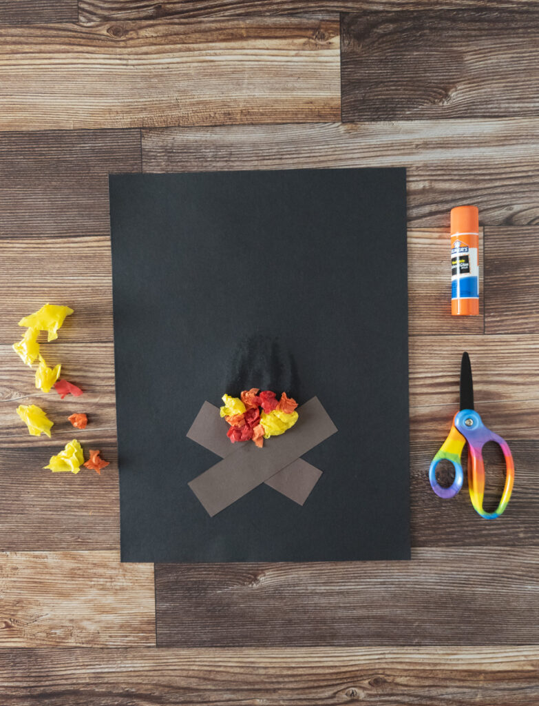 Ripped pieces of yellow, red, and orange tissue paper scrunched up and laying next to craft project. Other crumpled pieces are glued above the brown paper pieces of wood on black to make a fire.