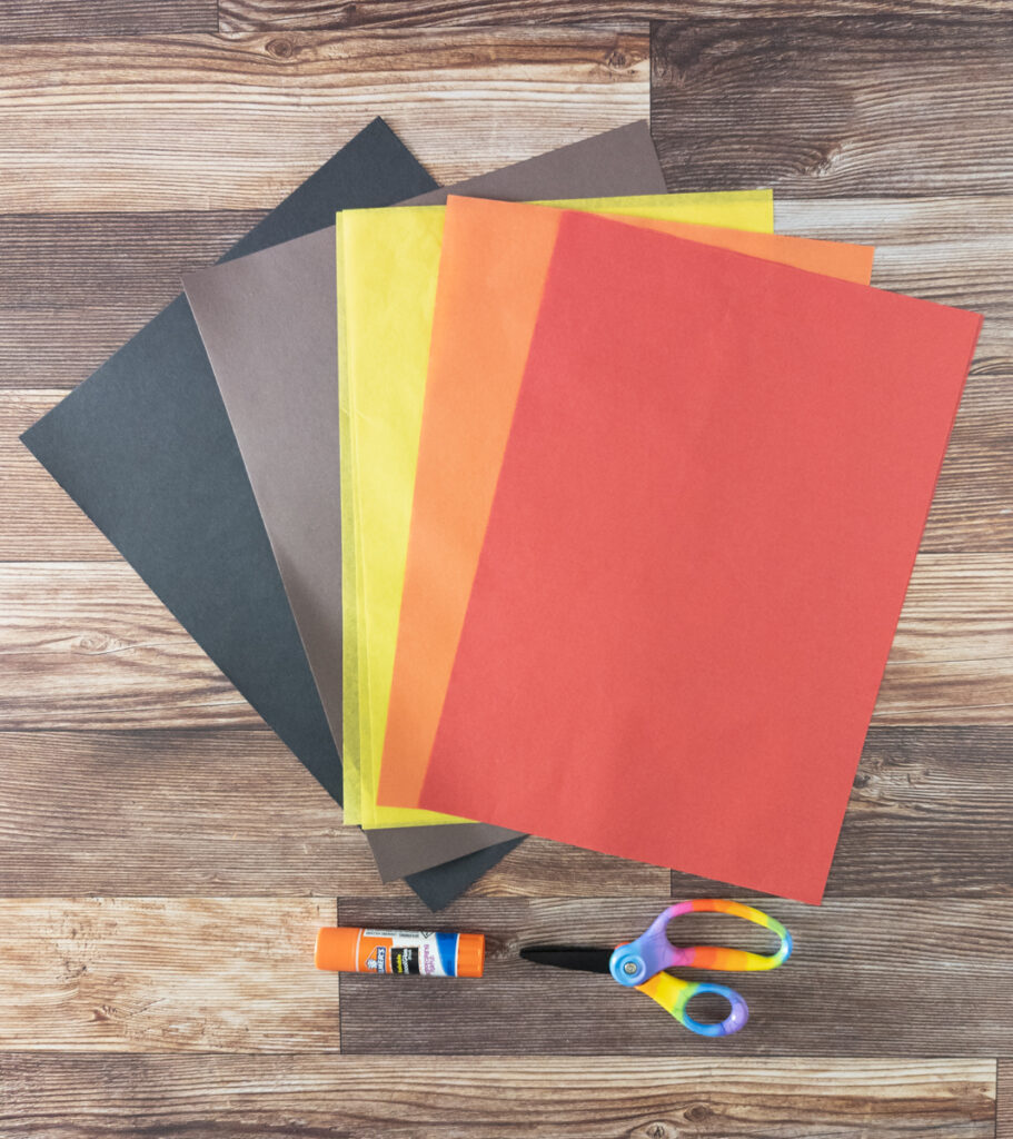 Black and brown construction paper and tissue paper in yellow, orange, and red are fanned out on a table above a glue stick and scissors.