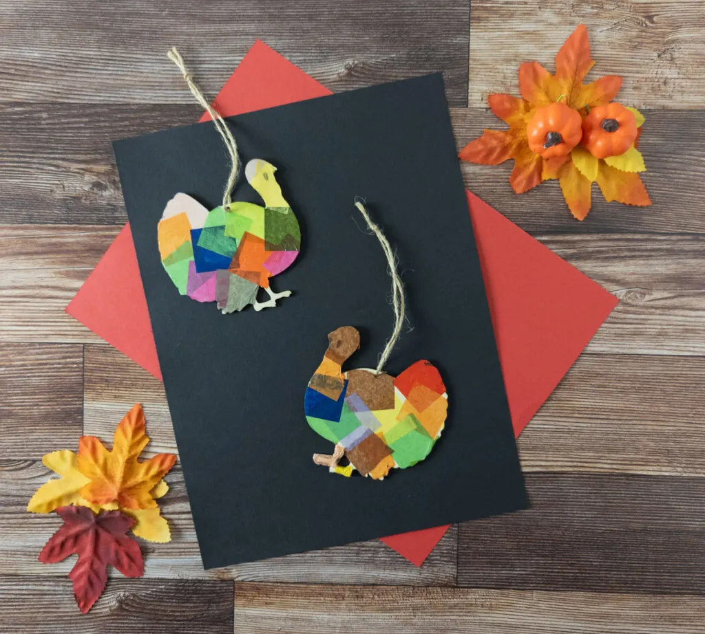 Top down view of two finished tissue paper turkey ornaments on black and red construction paper. Decorative fall leaves and small pumpkins are in the corners.
