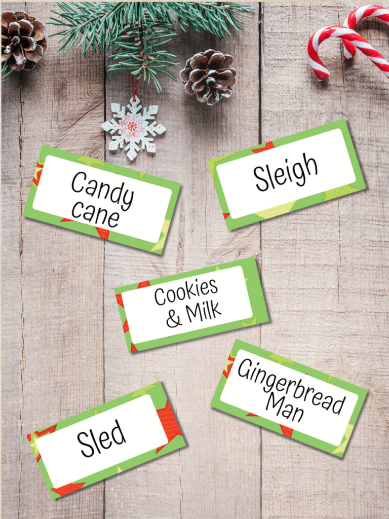 Mockup of five Christmas word cards for playing Pictionary on a festive background. The cards say candy cane, sleigh, cookies & milk, sled, and gingerbread man.
