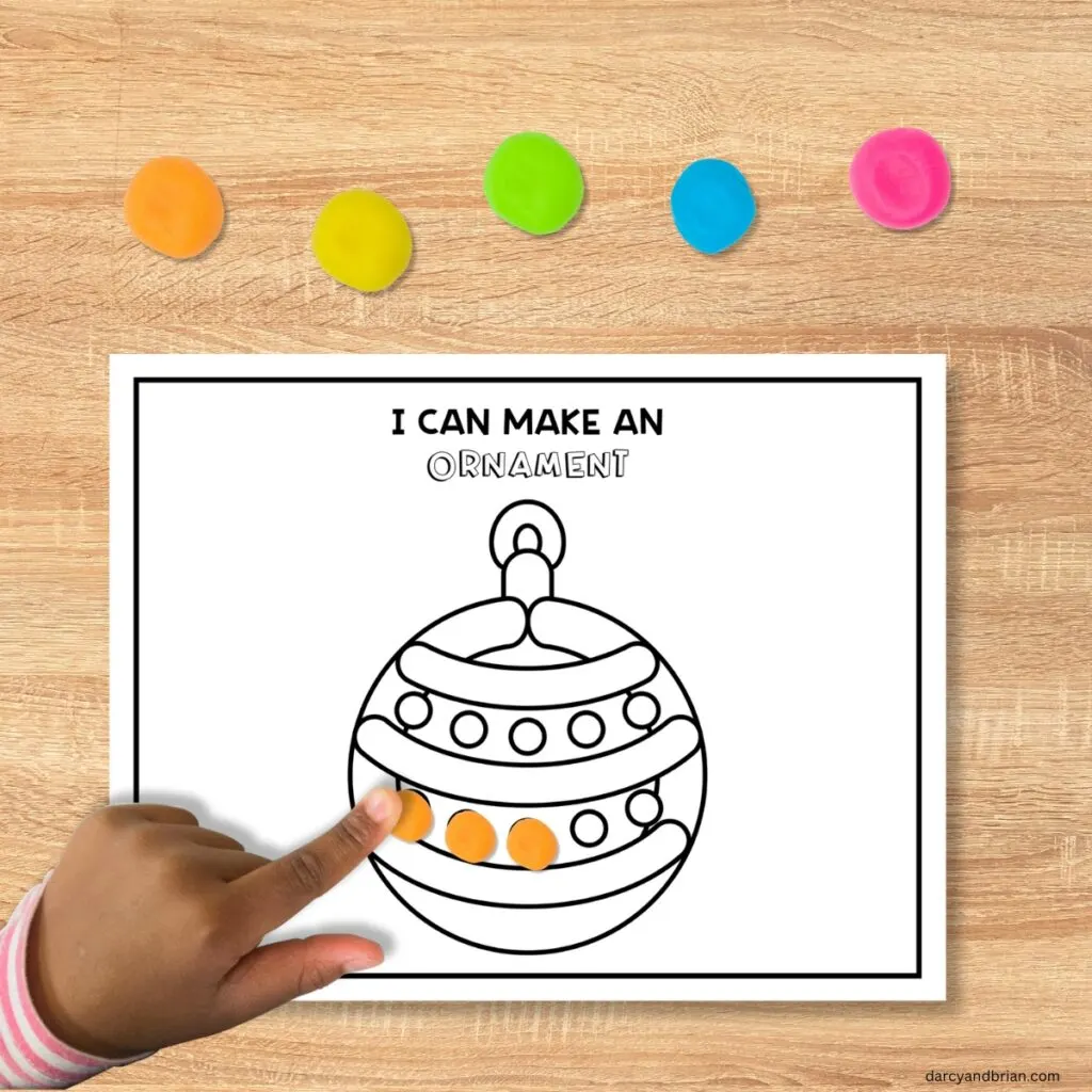 Child's hand with dark skin tone pressing small orange ball of playdough onto black and white version of an ornament playdough mat. Assorted colors of playdough balls along the top.