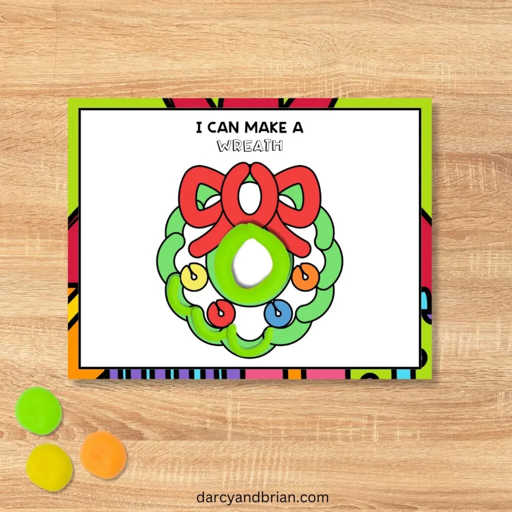 Mockup of full color version of the Christmas wreath playdough mat with pieces of green playdough on it. Green, orange, and yellow balls of dough in the lower left corner.